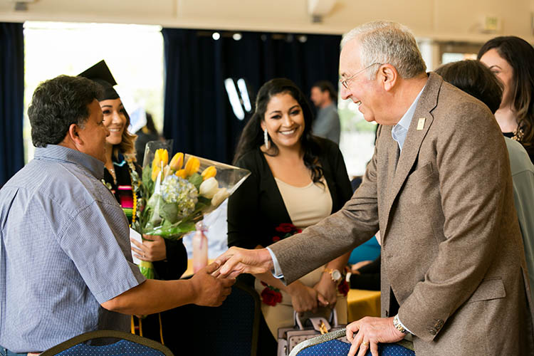 Robert Haas shakes the hand of a man while a woman in a cap and gown looks on