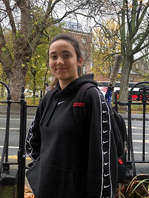 A girl wearing a dark hoodie stands in front of a fence, a street with traffic, and trees.