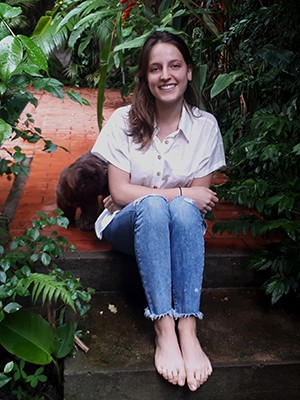 Isadora Saslavsky Muszkat at home in her garden in São Paulo. A girl wearing a white shirt and jeans sits among greenery.